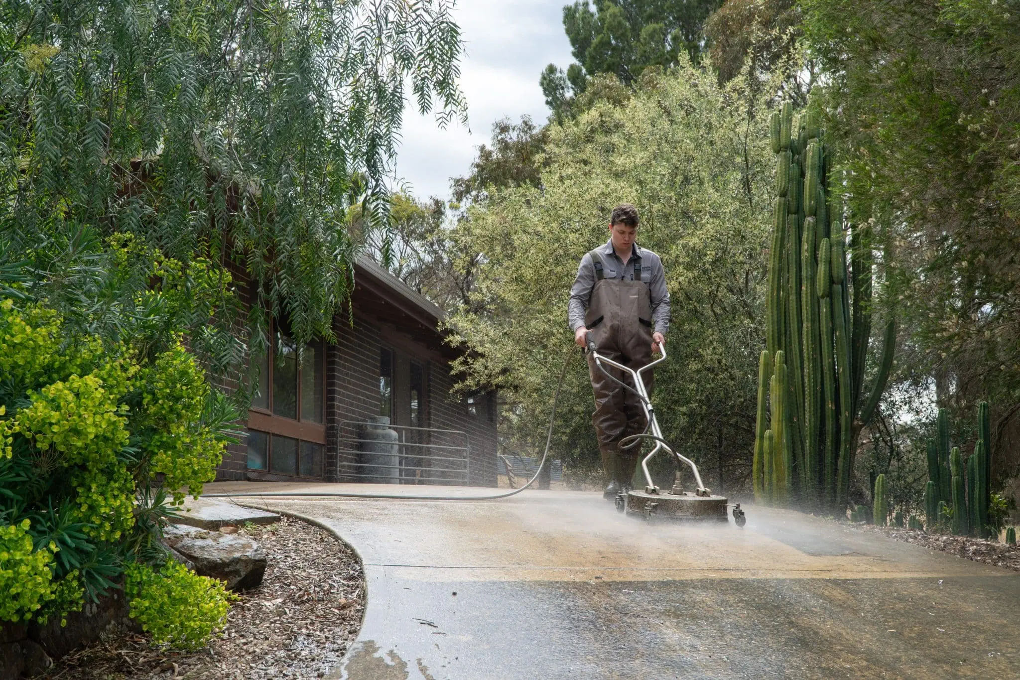 Technician performing Residential Pressure Cleaning in Adelaide, using high-pressure water equipment to clean an outdoor surface surrounded by greenery.